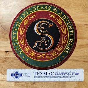 May include: A round, green and red embroidered patch with gold accents. The patch features a black circle with a white and gold stylized letter "S" and the text "SOCIETY of EXPLORERS & ADVENTURERS".