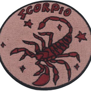 May include: A circular patch featuring a burgundy scorpion and the word "SCORPIO" in burgundy lettering. The patch has a light pink background with three stars and a diamond shape. The patch is outlined in black.