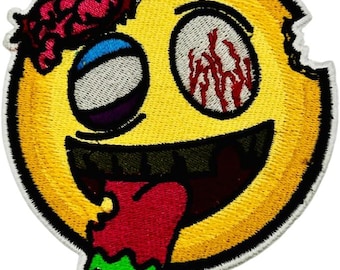 May include: A circular embroidered patch featuring a yellow zombie emoji with a missing chunk on the right side. It has one blue eye, one bloodshot eye, exposed pink brain matter on the top left, and a wide mouth with a red tongue dripping green slime.