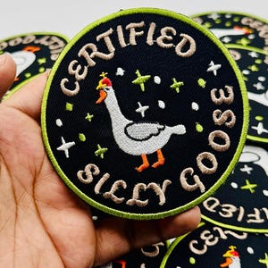May include: A round, black patch with the words "CERTIFIED SILLY GOOSE" embroidered in cream-colored thread. The patch features a cartoon goose wearing a crown, surrounded by stars and green accents. The patch is held in a hand.