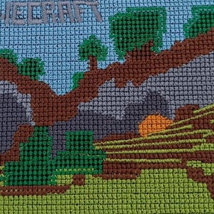 May include: A pixelated cross-stitch design featuring the word "MINECRAFT" at the top. The scene depicts a landscape with trees, a mountain, and a setting sun. The color palette includes shades of blue, green, brown, gray, and orange.