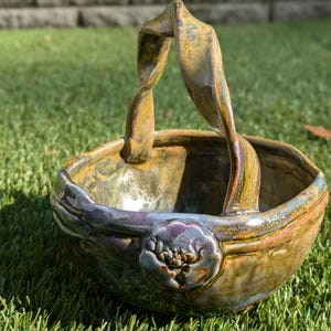 May include: A ceramic bowl with a twisted handle and a floral design. The bowl is glazed in shades of brown, gold, and purple.