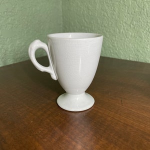 May include: A white ceramic teacup with a pedestal base and a curved handle. The cup has a slightly flared shape and a crackled surface texture. The handle is thick and rounded, and the cup is set on a dark wooden surface.