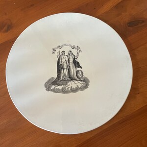 May include: A white, round plate with a black and white illustration of two figures holding a scale and a sword, with the text "Justitia et Fides" on a banner above them. The plate has a simple, elegant design.