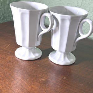 May include: Two white, pedestal-style coffee cups with angular sides and loop handles. The cups sit on a wooden surface, and the background is a light green wall. The cups are likely made of ceramic or porcelain.