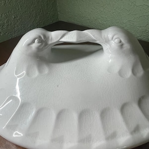May include: White ceramic serving dish with a handle shaped like two bird heads. The dish has a scalloped edge and a textured surface.