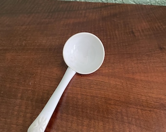 May include: A white ceramic ladle with a rounded bowl and a long handle. The handle has a decorative design.