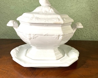 May include: White porcelain soup tureen with a lid and handles, resting on a matching octagonal plate. The tureen features embossed floral detailing and a decorative finial on the lid. A classic piece for serving soups or stews.