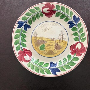 May include: A round, cream-colored decorative plate with a painted scene of rabbits in a field. The plate's rim features a floral design with red, blue, and green accents. The plate is approximately 10 inches in diameter.