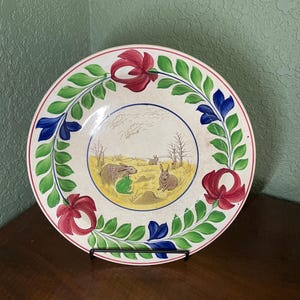 May include: A vintage ceramic plate with a hand-painted design of rabbits in a field. The plate has a white background with a yellow field, green leaves, blue flowers, and red flowers around the edge.