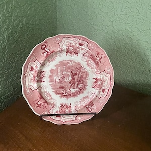 May include: A pink and white ceramic plate with a scalloped edge. The plate features a detailed floral design and a central scene depicting a man and a woman in a garden setting.