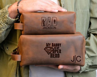 May include: Two brown leather toiletry bags. The top bag has the word "DAD" with a silhouette of a person fishing and the name "JACKSON". The bottom bag says "MY DADDY IS MY SUPER HERO" with a Superman logo and the initials "J|C".
