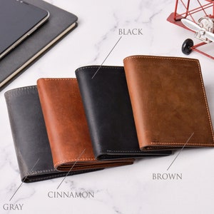 May include: Four leather wallets in grey, cinnamon, black, and brown are displayed. The wallets are closed, showing the exterior with visible stitching. The wallets are arranged in a row, with the colours labelled.