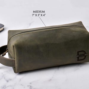 May include: A medium-sized, olive green leather toiletry bag with a zipper closure and a handle. The bag is rectangular, measuring 7" x 3" x 4". The bag has the word "DAVID" and the letter "D" engraved on the front.