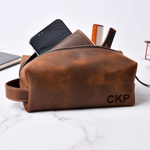 May include: A brown leather toiletry bag with a zipper closure, handle, and the initials "CKP" embossed on the front. The bag is open, revealing a smartphone, wallet, and two combs. The bag is designed for travel and personal care items.