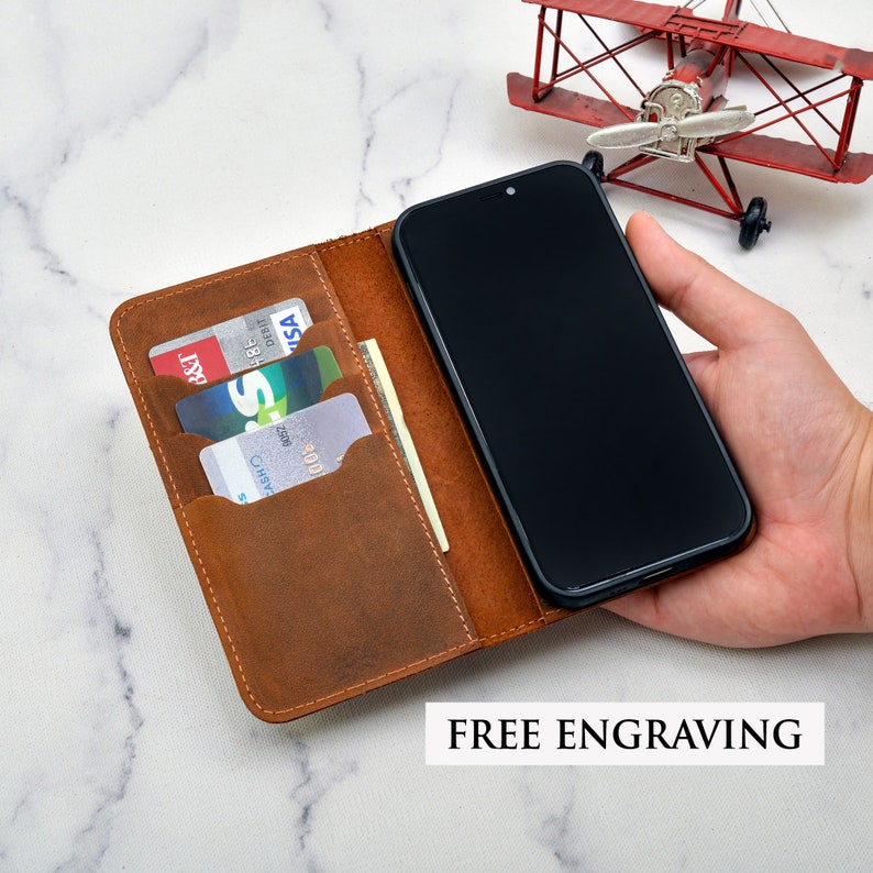 May include: A brown leather phone case with slots for credit cards. The case is open and a black smartphone is visible. The text "FREE ENGRAVING" is printed below the case.