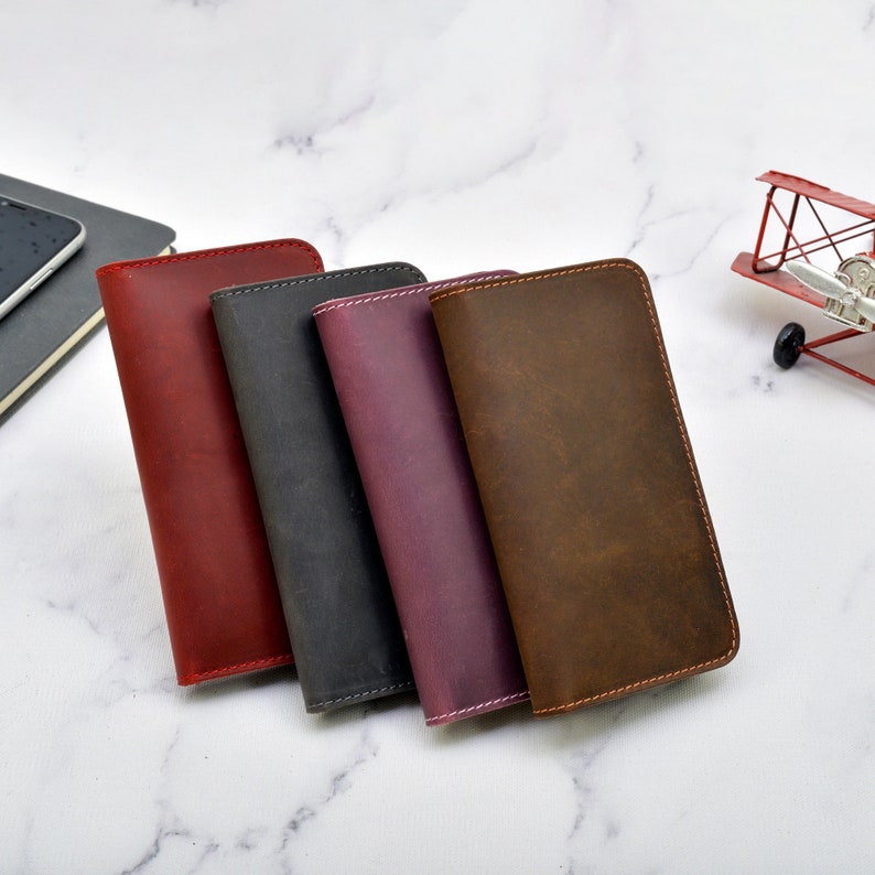 May include: Four leather wallets in different colours: red, black, purple, and brown. The wallets are stacked on top of each other and have a stitched edge.