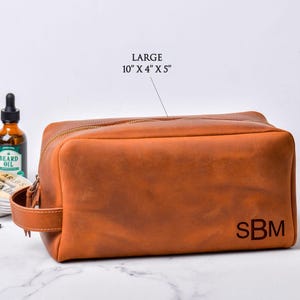 May include: A large, brown leather toiletry bag with a zipper closure and a handle. The bag is embossed with the initials "SBM" and measures 10" x 4" x 5". A bottle of beard oil is visible in the background.