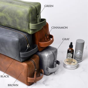 May include: A stack of leather toiletry bags in various colors: green, cinnamon, gray, and brown. Each bag has a zipper closure and a handle. Grooming products, including beard oil and pomade, are displayed next to the bags.