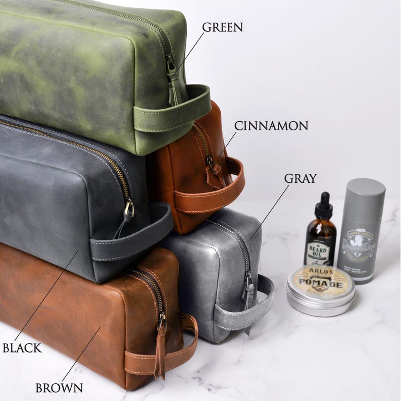 May include: A stack of leather toiletry bags in various colors: green, cinnamon, gray, and brown. Each bag has a zipper closure and a handle. Grooming products, including beard oil and pomade, are displayed next to the bags.