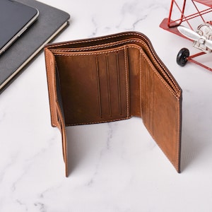 May include: Open brown leather wallet with several card slots and a bill compartment. The wallet is made of textured leather and has white stitching around the edges. The wallet is open, showing its interior.