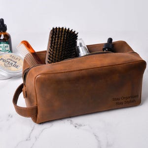 May include: A brown leather toiletry bag with a zipper closure, handle, and the text "Stay Organized Stay Stylish." The bag contains a comb, brush, and grooming products. A bottle of beard oil and pomade are visible.