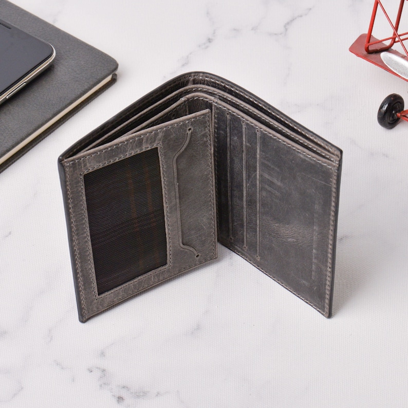 May include: Open grey leather wallet with multiple card slots and a clear ID window. The wallet is displayed open, showcasing its interior design. The wallet is on a white surface.