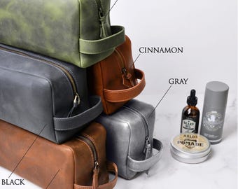 May include: A selection of leather wash bags in shades of green, cinnamon, grey, and brown. Each bag features a zip closure and a handle. Grooming products, such as beard oil and pomade, are arranged beside the bags.