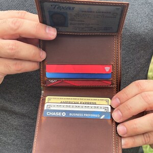 May include: Open brown leather wallet with multiple card slots, holding credit cards and a driver's license. The wallet is held open, showcasing its interior design and the cards within. The stitching is visible.
