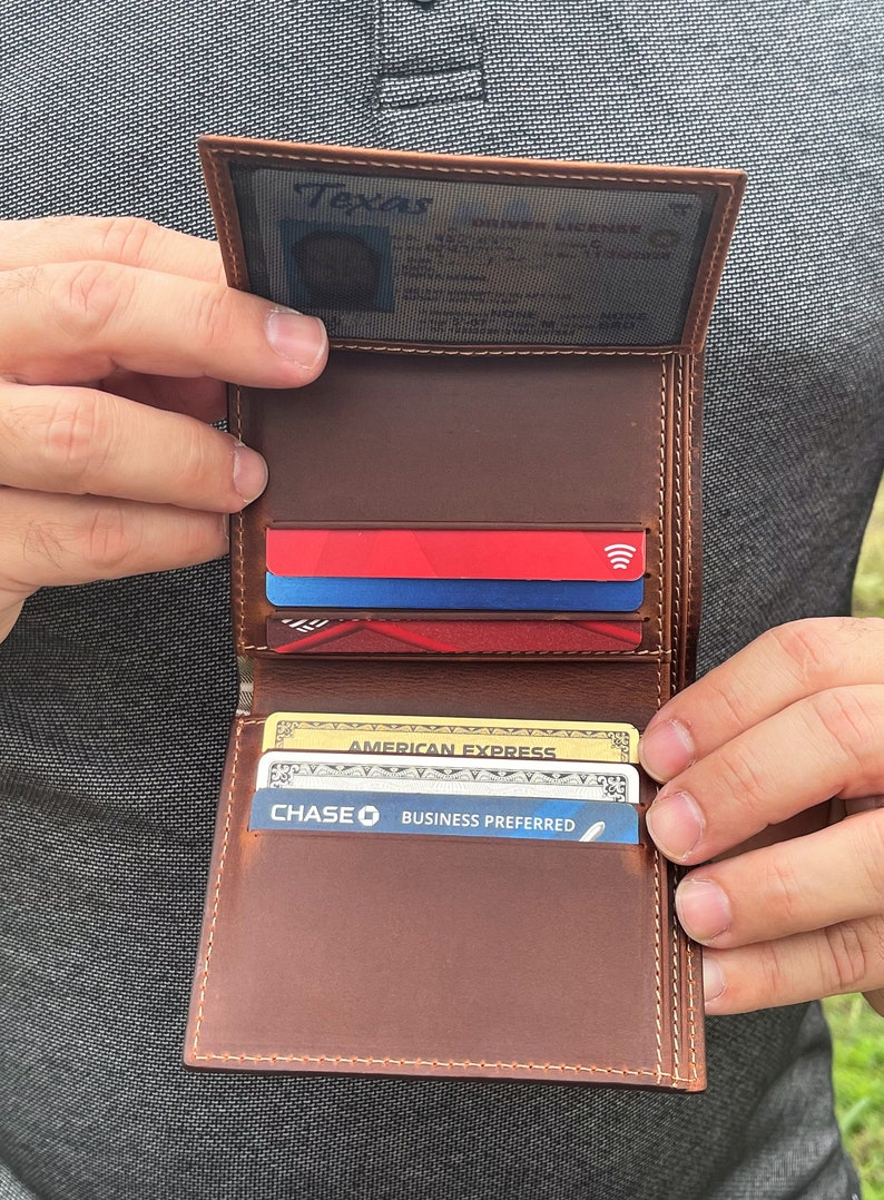 May include: Open brown leather wallet with multiple card slots, holding credit cards and a driver's license. The wallet is held open, showcasing its interior design and the cards within. The stitching is visible.