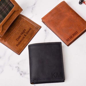 May include: Three leather wallets in shades of brown and black are displayed. One is open, revealing a plaid interior and the text "Date Night Rebecca & David." Another has the date "02.02.2022" embossed. The third wallet has the initials "KM."
