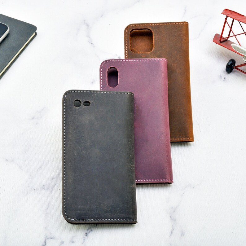May include: Three leather phone cases in different colours: dark grey, purple, and brown. The cases are closed and have a stitched edge.