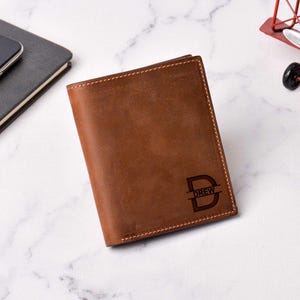 May include: A brown leather wallet with the name "DREW" and a stylized "D" engraved on the front. The wallet is closed and has visible stitching around the edges. The background is a marble surface.