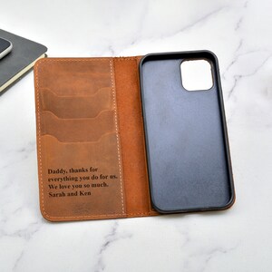 May include: A brown leather phone case with a wallet flap. The case has a personalised message that reads "Daddy, thanks for everything you do for us. We love you so much. Sarah and Ken".