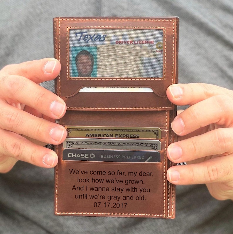 May include: A brown leather wallet, open to reveal a Texas driver's license in a clear window. Several credit cards are visible. The wallet is engraved with the message: "We've come so far, my dear, look how we've grown." The date 07.17.2017 is also engraved.