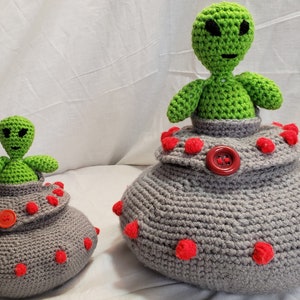 May include: Two crocheted alien toys in spaceships. The larger spaceship is gray with red accents and the smaller spaceship is gray with red accents. Both spaceships have a crocheted alien inside.
