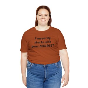May include: A rust-colored t-shirt with short sleeves and a crew neck. The shirt has the text "Prosperity starts with your MINDSET" printed in black.