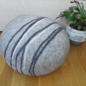 May include: A round, gray and white felted pouf with a striped pattern. The pouf is sitting on a wooden floor.