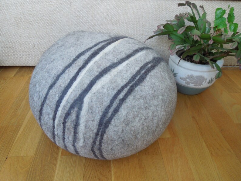 May include: A round, gray and white felted pouf with a striped pattern. The pouf is sitting on a wooden floor.