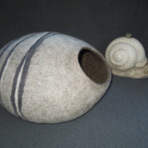 May include: A gray and white felted wool cat bed shaped like a half sphere with a round opening. The bed has black stripes. A white ceramic snail figurine is next to the bed.
