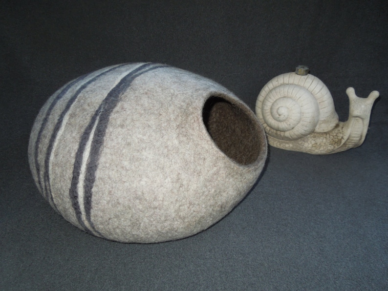 May include: A gray and white felted wool cat bed shaped like a half sphere with a round opening. The bed has black stripes. A white ceramic snail figurine is next to the bed.