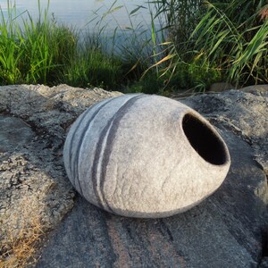 May include: A gray and white felted cat bed shaped like a large oval stone with a black opening. The bed is sitting on a rocky surface.