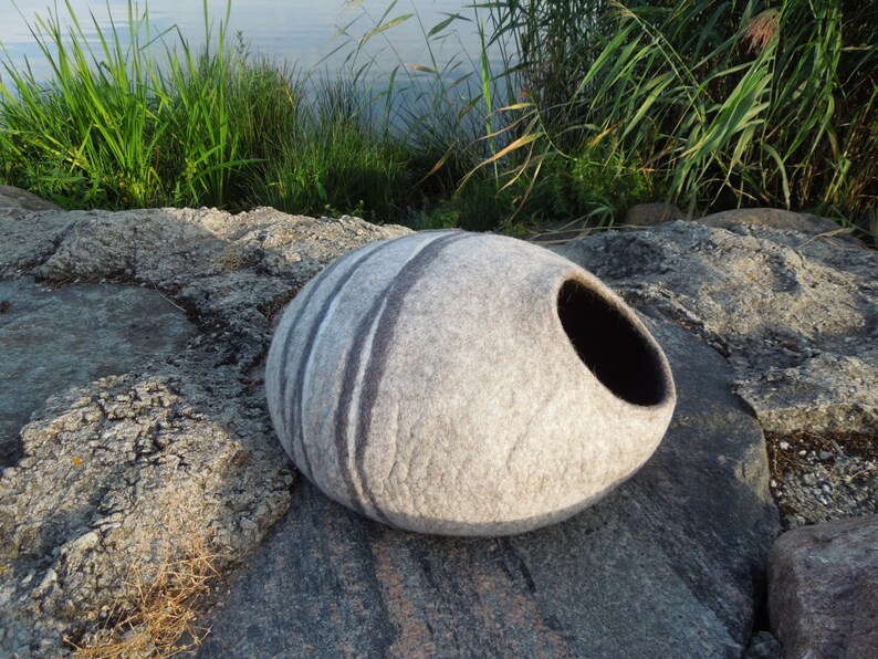 May include: A gray and white felted cat bed shaped like a large oval stone with a black opening. The bed is sitting on a rocky surface.