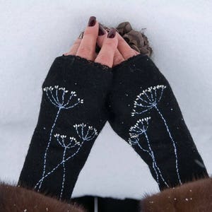 Pu&ograve; includere: Guanti senza dita neri con ricami floreali bianchi. I guanti sono realizzati in tessuto scuro e presentano un motivo delicato e dettagliato di fiori stilizzati. I guanti sono mostrati su uno sfondo innevato.