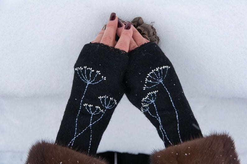 Pu&ograve; includere: Guanti senza dita neri con ricami floreali bianchi. I guanti sono realizzati in tessuto scuro e presentano un motivo delicato e dettagliato di fiori stilizzati. I guanti sono mostrati su uno sfondo innevato.