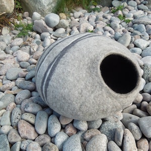 May include: A gray felt cat bed shaped like a rounded stone with a black felt entrance hole. The bed is sitting on a bed of gray and white stones.