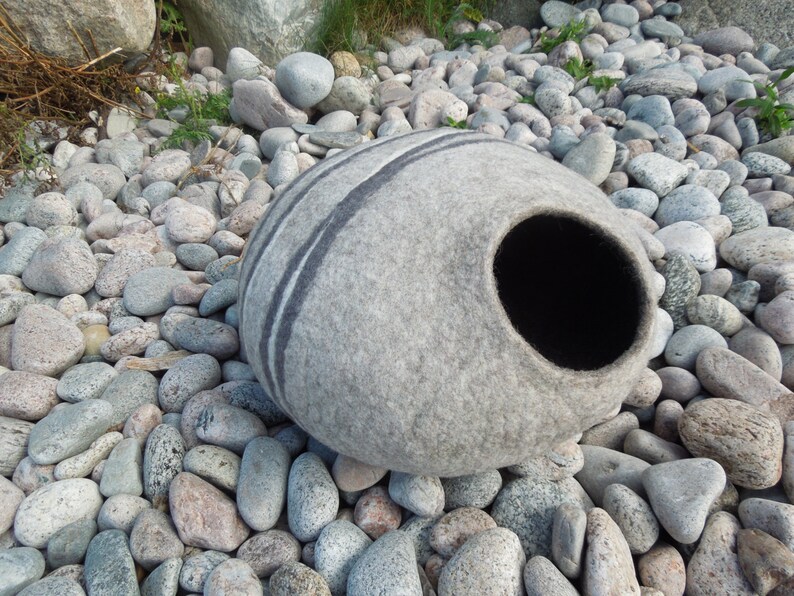 May include: A gray felt cat bed shaped like a rounded stone with a black felt entrance hole. The bed is sitting on a bed of gray and white stones.