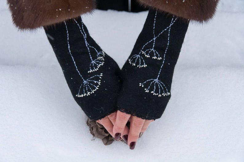 Pu&ograve; includere: Guanti neri senza dita con ricami floreali bianchi. I guanti sono realizzati in un materiale scuro e presentano un motivo decorativo di delicate cuciture bianche. I guanti sono mostrati su uno sfondo innevato.