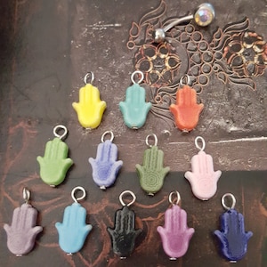 May include: A collection of colorful Hamsa hand charms in various shades including yellow, green, blue, orange, pink, purple, and black. Each charm has a silver loop for attachment. A belly button ring is also visible.