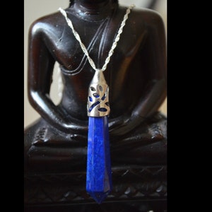 May include: A silver pendant necklace featuring a faceted, deep blue gemstone. The pendant has a silver cap with a decorative cutout design. The necklace is displayed against a dark background.